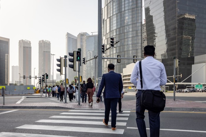 People crossing a street at a crosswalk in Dubai's Business Bay financial district, with modern skyscrapers in the background.